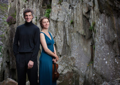 two musicians stand side by side one holds a violin surrounded by rock by Anglesey commercial photographer Gill Jones Photography