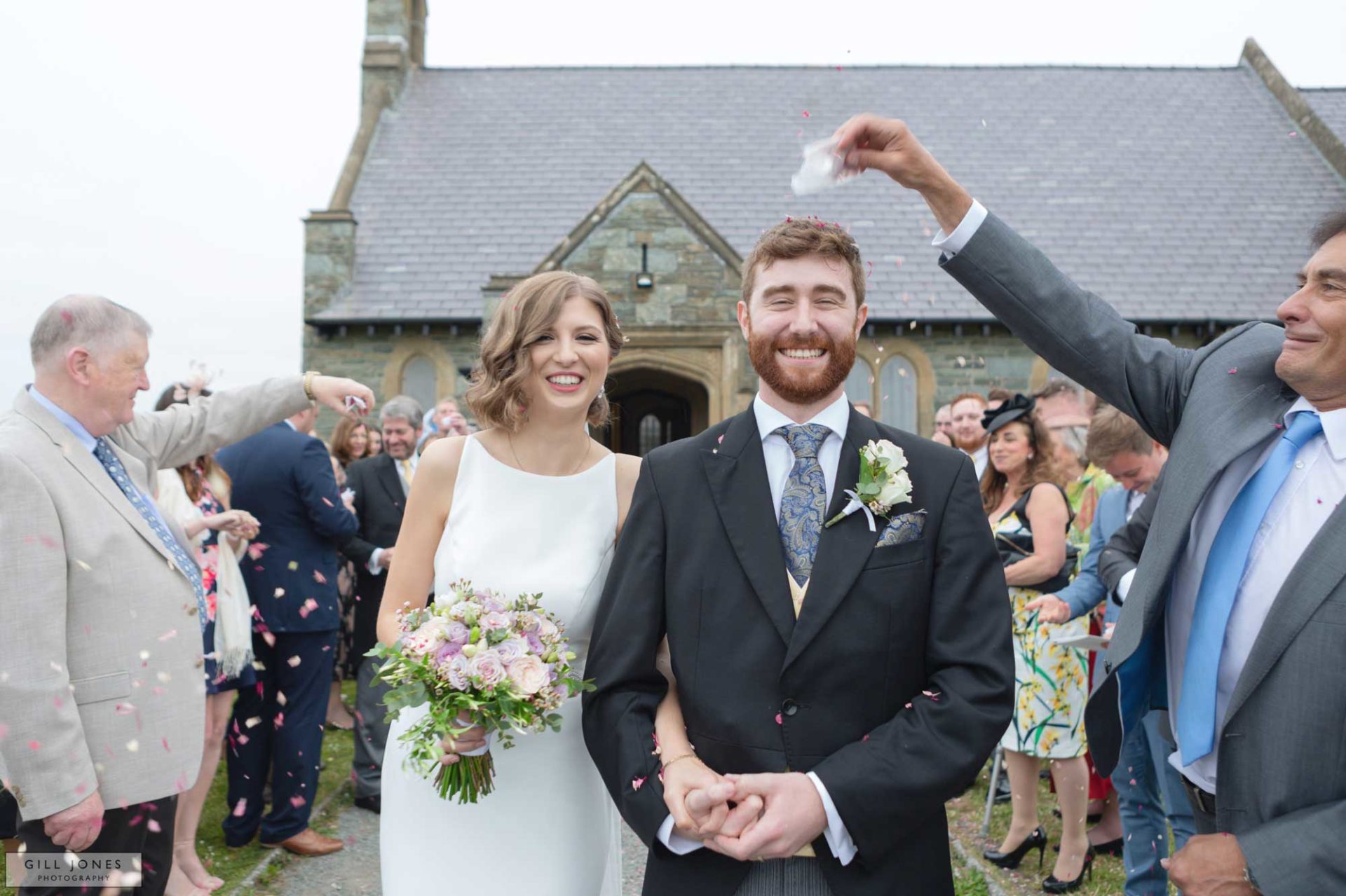 confetti is being shaken on top of the grooms head outside of St. Ffraids church in Trearddur Bay