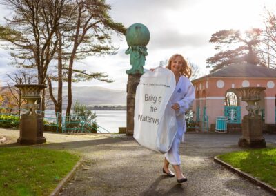 in the bright morning sunshine a bridesmaid carries a wedding dress enclosed in a cover through Portmeirion Village by Anglesey wedding photographer Gill Jones Photography