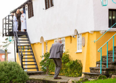 a male wedding guest looks up a flight of stairs towards the bride and two other female friends they all laugh by Anglesey wedding photographer Gill Jones Photography