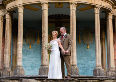 a bride and groom stand in front of a semicircle of columns the groom holds his bride's little finger at Portmeirion Village by Anglesey wedding photographer Gill Jones Photography