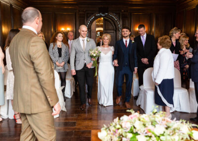 a bride walks down the aisle towards her groom flanked by two young men in Hercules Hall at Portmeirion by Anglesey wedding photographer Gill Jones Photography