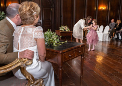a young bridesmaid takes a photograph of a bride and groom as they sit behind the register desk in Hercules Hall at Portmeirion Village by Anglesey wedding photographer Gill Jones Photography