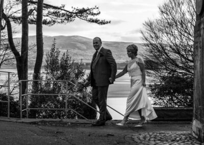 a groom leads his bride by her hand whilst holding her bouquet the Dwyryd estuary is in the background by Anglesey wedding photographer Gill Jones Photography