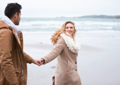 couple walking hand in hand, the woman leads the man, towards Llanddwyn island by Anglesey wedding photographer Gill Jones