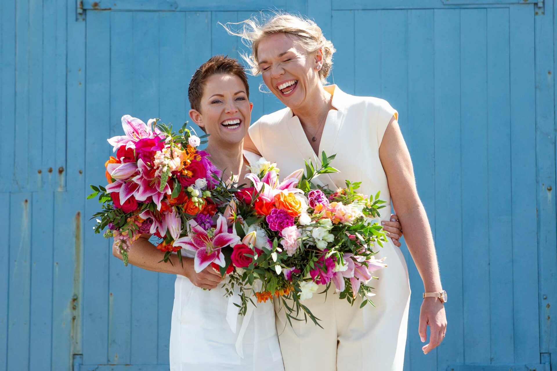 Couple standing infront of an old blue lifeboat hut door , laughing and holding their colourful bouquets by Anglesey wedding Photographer Gill Jones