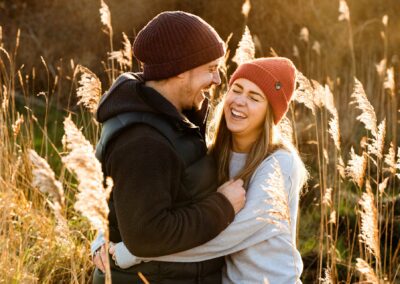 Couple hugging and laughing surrounded by seed heads as the sun drops by Anglesey wedding Photographer Gill Jones