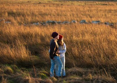 Couple standing in field of straw looking towards the sun as it sets by Anglesey wedding Photographer Gill Jones