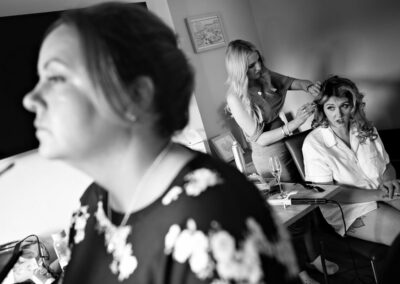 the bride has her hair done in the backgrounds as her bridesmaid has her makeup done in the foreground by Anglesey wedding photographer, North Wales, Gill Jones Photography