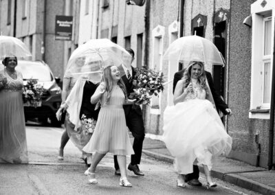the bride and bride's party walk towards the church holding umbrellas by Anglesey wedding photographer, North Wales, Gill Jones Photography