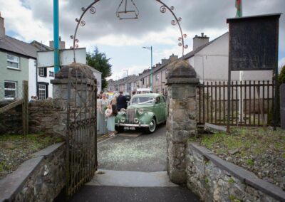 church path leads to wedding car with wedding guests looking on by Anglesey wedding photographer, North Wales, Gill Jones Photography