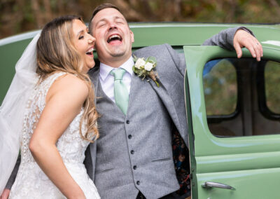 bride and groom laugh out loud, throwing their heads back, leaning against a car door by Anglesey wedding photographer, North Wales, Gill Jones Photography