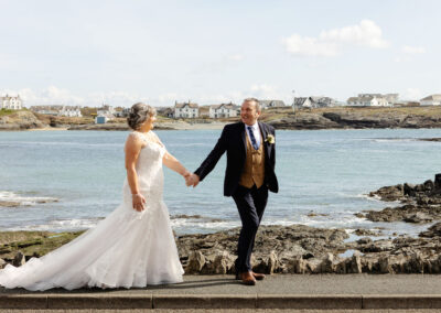 groom leads his new wife along the pavement with the beach in the background