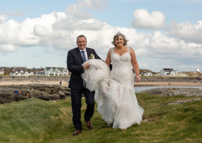 bride and groom walking on the headland in trearddur bay, groom holds his wife's train