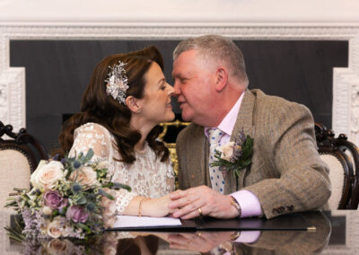bride and groom sitting behind desk leaning towards one another to kiss by Anglesey wedding photographer Gill Jones Photography