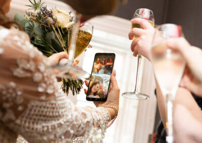bride holds mobile phone and talks to her family, champagne in hand by Anglesey wedding photographer Gill Jones Photography