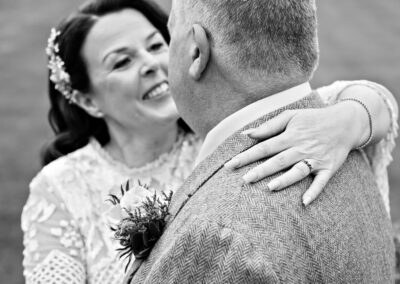 bride's engagement ring on hand in foreground as bride smiles towards her groom by Anglesey wedding photographer Gill Jones Photography