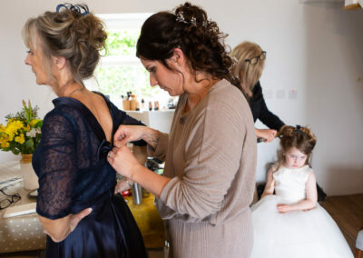 the bride pins a ribbon to the back of her mother's dress whilst her daughter has her hair styled by Anglesey photographer Gill Jones Photography