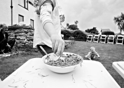 a girl brings grass to a pudding bowl whilst her Grandma watches her little sister crawl by Anglesey photographer Gill Jones Photography