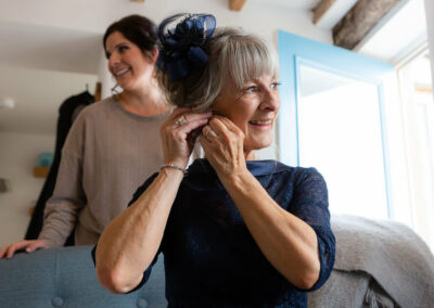 a mother of the bride attaches an earing to her ear as her daughter looks on in the background resting on a settee by Anglesey photographer Gill Jones Photography