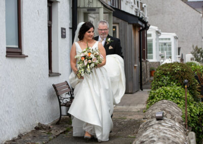 a bride walks towards an aisle in a garden as her father holds the train of her dress by Anglesey photographer Gill Jones Photography