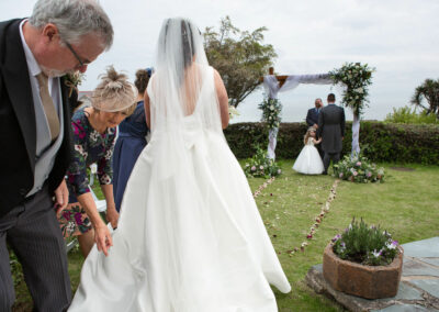 the bride begins her walk towards her groom as her father and mother in law perfect her dress by Anglesey photographer Gill Jones Photography