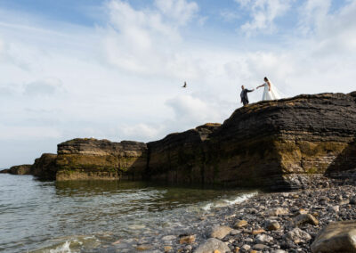 a groom offers his hand to his bride to guide her along the top of a cliff as a bird flies by by Anglesey photographer Gill Jones Photography