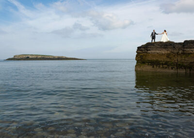 the bride and groom reach their hands to touch as they stand on top of the rocks with the sea below by Anglesey photographer Gill Jones Photography