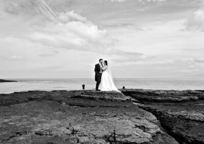 the bride and groom face each other standing on the rocks with fishermen in the background by Anglesey photographer Gill Jones Photography