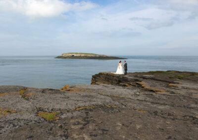 Moelfre Island is in the background as a bride and groom satnd in the foreground on rocks by Anglesey photographer Gill Jones Photography