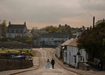 a bride and groom walk down the hill in the centre of the road in the seaside town of Moelfre by Anglesey photographer Gill Jones Photography