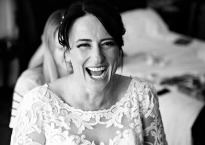 bride smiles a huge smile as her friend buttons her wedding dress from behind by Anglesey wedding photographer Gill Jones Photography