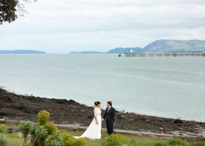 bride and groom hand in hand standing on the edge of the coast line on the Menai Straits by Anglesey wedding photographer Gill Jones Photography