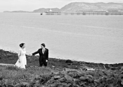 groom guiding bride along the shoreline with Bangor Pier in the background by Anglesey wedding photographer Gill Jones Photography