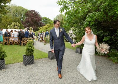 couple walking hand in hand after their wedding ceremony, guests in the background, the bride is smiling widely by Anglesey wedding photographer Gill Jones