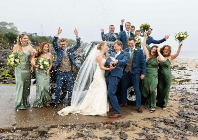 A bride and groom stand by a Landrover vehicle on Traeth Bychan beach with their wedding party, the groom opens a bottle of champagne and sprays it everywhere by Anglesey Photographer Gill Jones Photography