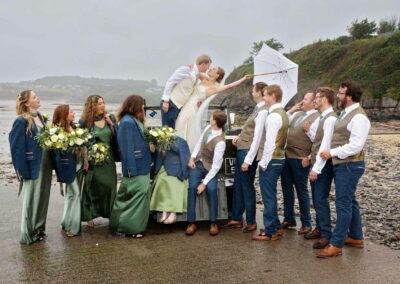 A bride and groom stand on the back of a Landrover vehicle and kiss one another, their wedding party cheers by Anglesey Photographer Gill Jones Photography