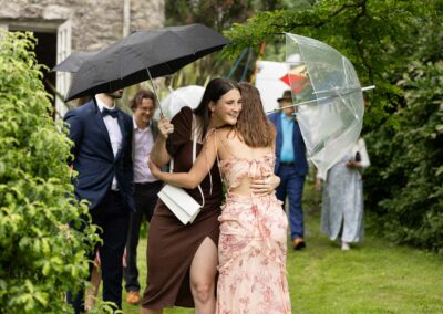 Wedding guests hug eachother holding umbrellas just to the side so as not to lock by Anglesey Photographer Gill Jones Photography