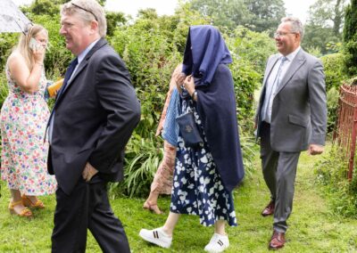 Wedding guests walk back and forth, one woman wears her scarf over her head to shelter from the rain by Anglesey Photographer Gill Jones Photography