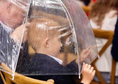 A young boy is sheltering from the rain and traces his finger on the inside of the umbrella as if writing in the raindrops by Anglesey Photographer Gill Jones Photography