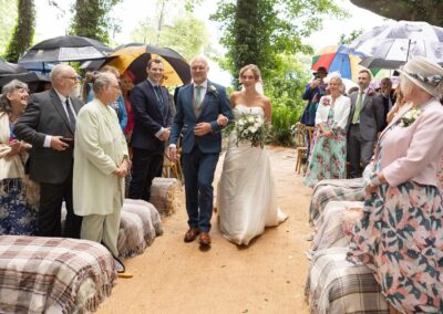 The bride walks towards her groom linking arms with her father, guests are standing holding umbrellas by Anglesey Photographer Gill Jones Photography