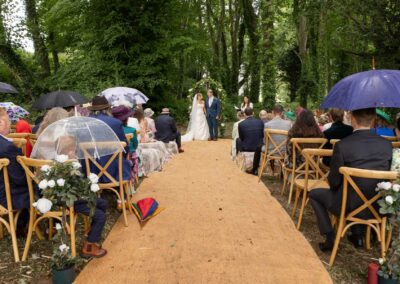 Bride and Groom stand together underneath an ivy covered arch, guets are seated left and right of the aisle holding umbrellas sheltering from the rain by Anglesey Photographer Gill Jones Photography