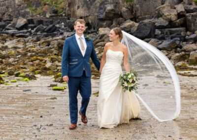Bride and Groom walking hand in hand along Traeth Bychan, they're smiling broadly as the bride looks towards the groom her veil blows in the wind by Anglesey Photographer Gill Jones Photography