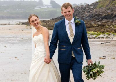 Bride and Groom walking hand in hand along Traeth Bychan, they're smiling broadly as the groom carries the bouquet by Anglesey Photographer Gill Jones Photography