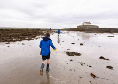 Two boys wearing blue anoraks run along the wet sand towards St. Cwyfan's Church by Anglesey Photographer Gill Jones Photography