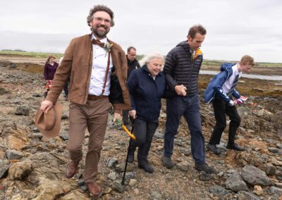 A Grooms leads his guests towards St. Cwyfan's Church, his mother leans on a walking stick whilst holding a man's hand by Anglesey Photographer Gill Jones Photography