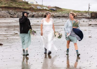 A bride strides forward with her two bridesmaids, the weather is wet and windy and they're wearing wellies by Anglesey Photographer Gill Jones Photography