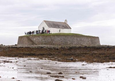 Guests await the arrival of the bride as they stand on the island at St. Cwyfan's which is behind the group by Anglesey Photographer Gill Jones Photography