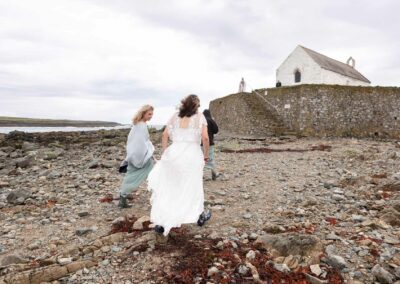 a bride walks with her two bridesmaids towards St. Cwyfan's Church, the Vicar stands at the top of the stairs to welcome them by Anglesey Photographer Gill Jones Photography
