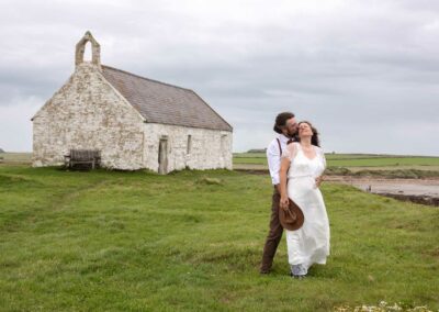 With St. Cwyfan's, The Church in the Sea in the background a bride and groom caress by Anglesey Photographer Gill Jones Photography